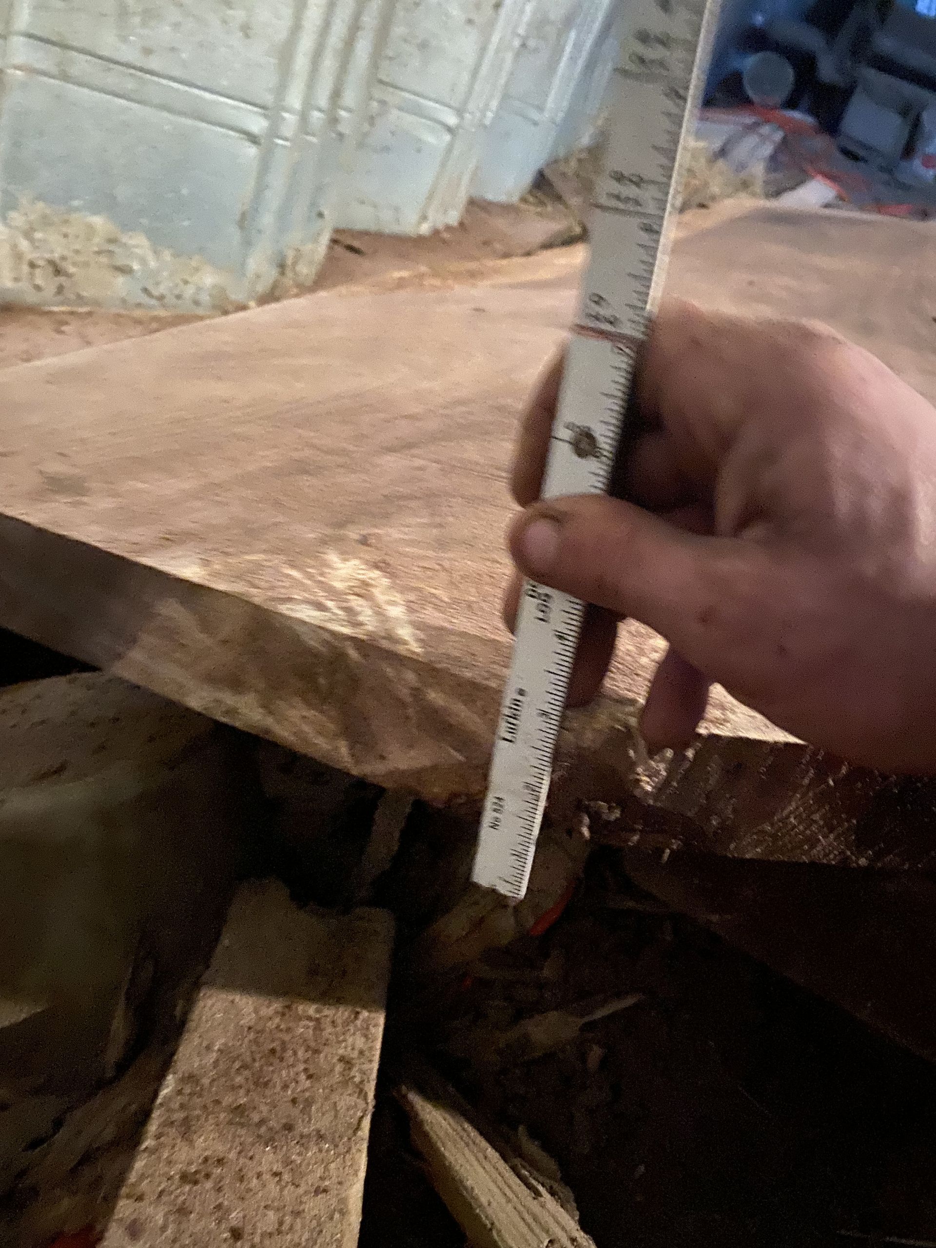 Hand holding ruler, measuring distance between wooden beams in a dim, earthy-toned space with metal sheeting.