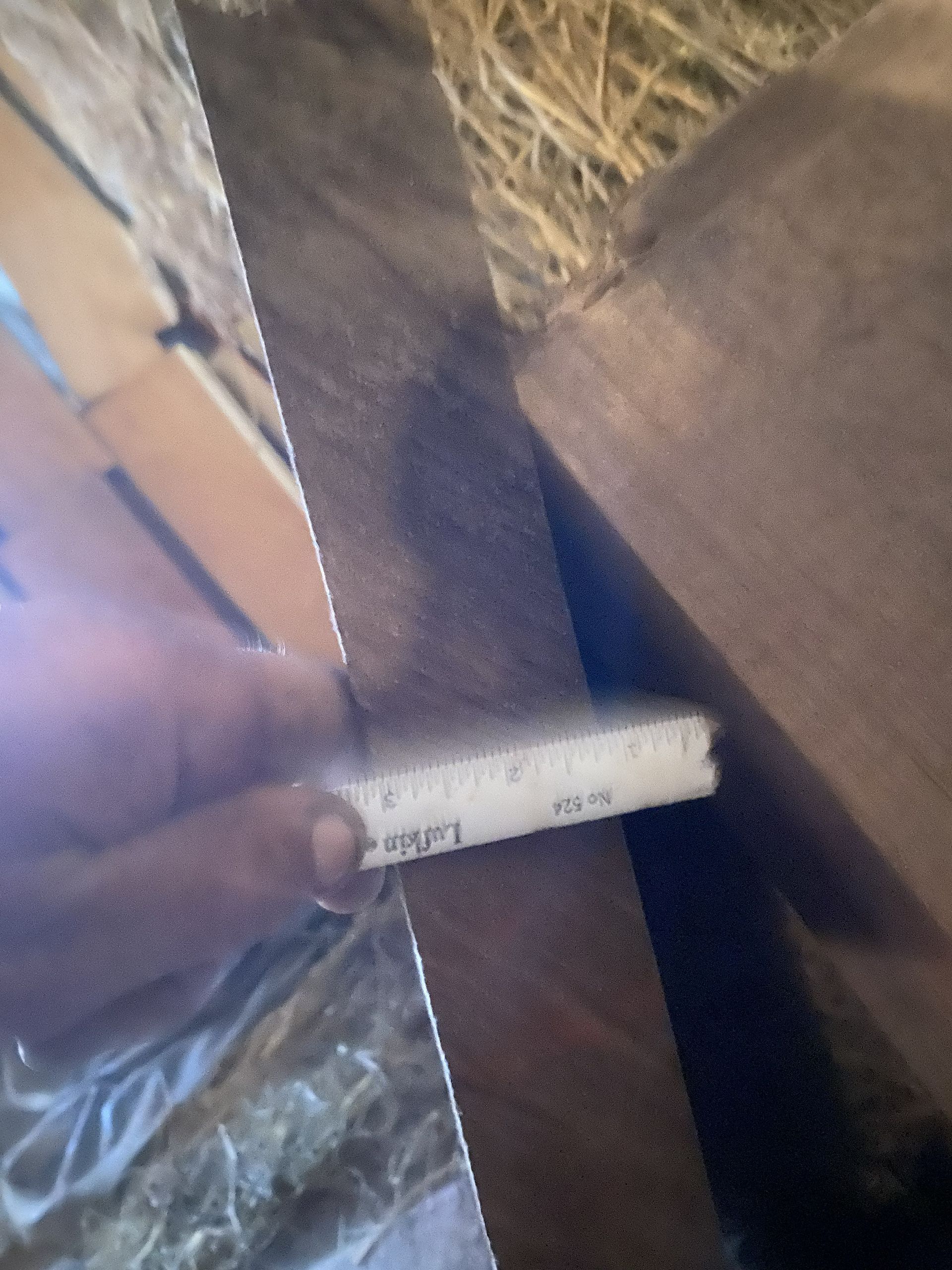 A person holds a white measuring tape near a dark brown wooden structure with hay in the background.
