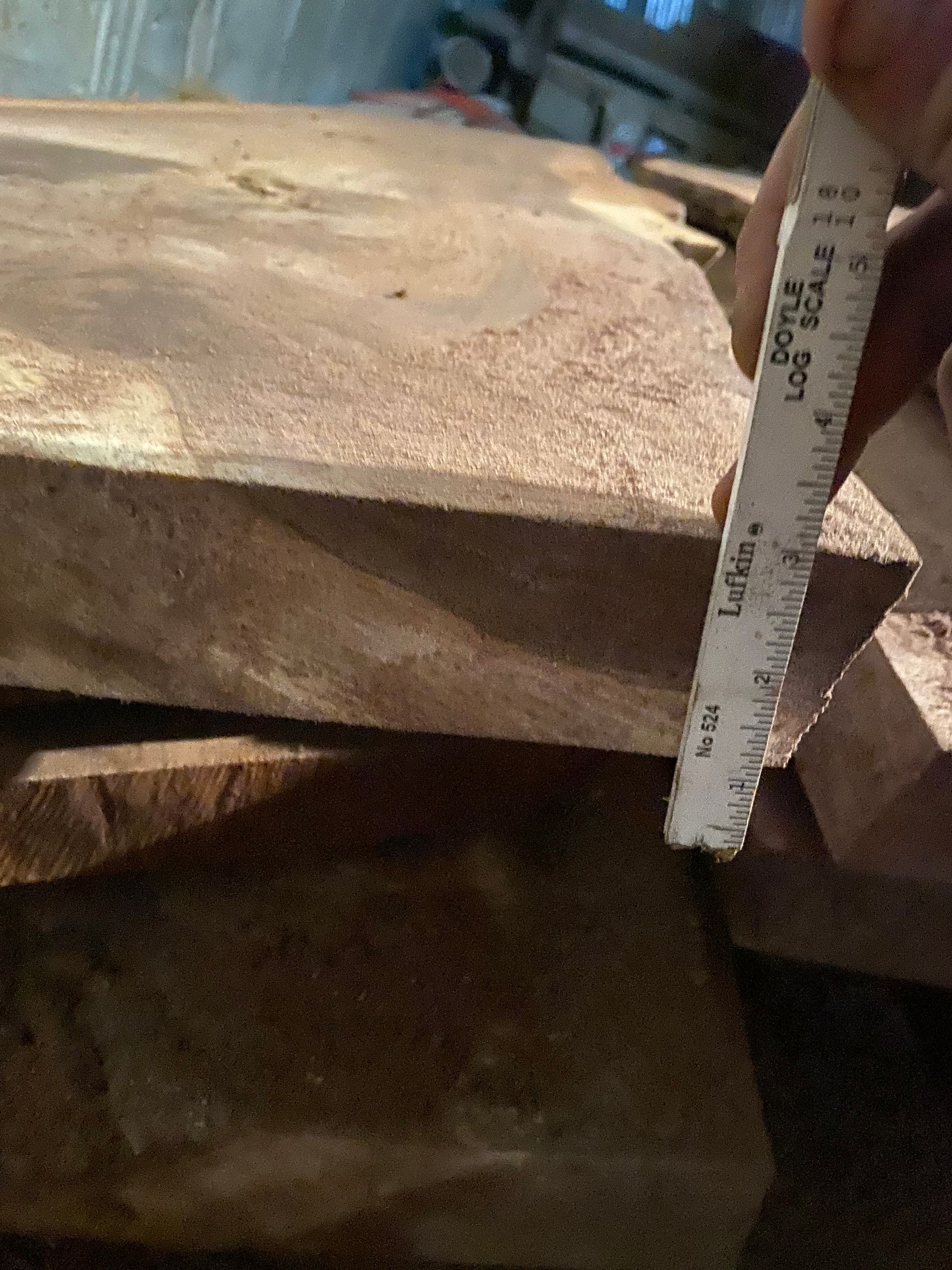 A hand measuring the thickness of a wooden plank with a log scale ruler in a workshop.