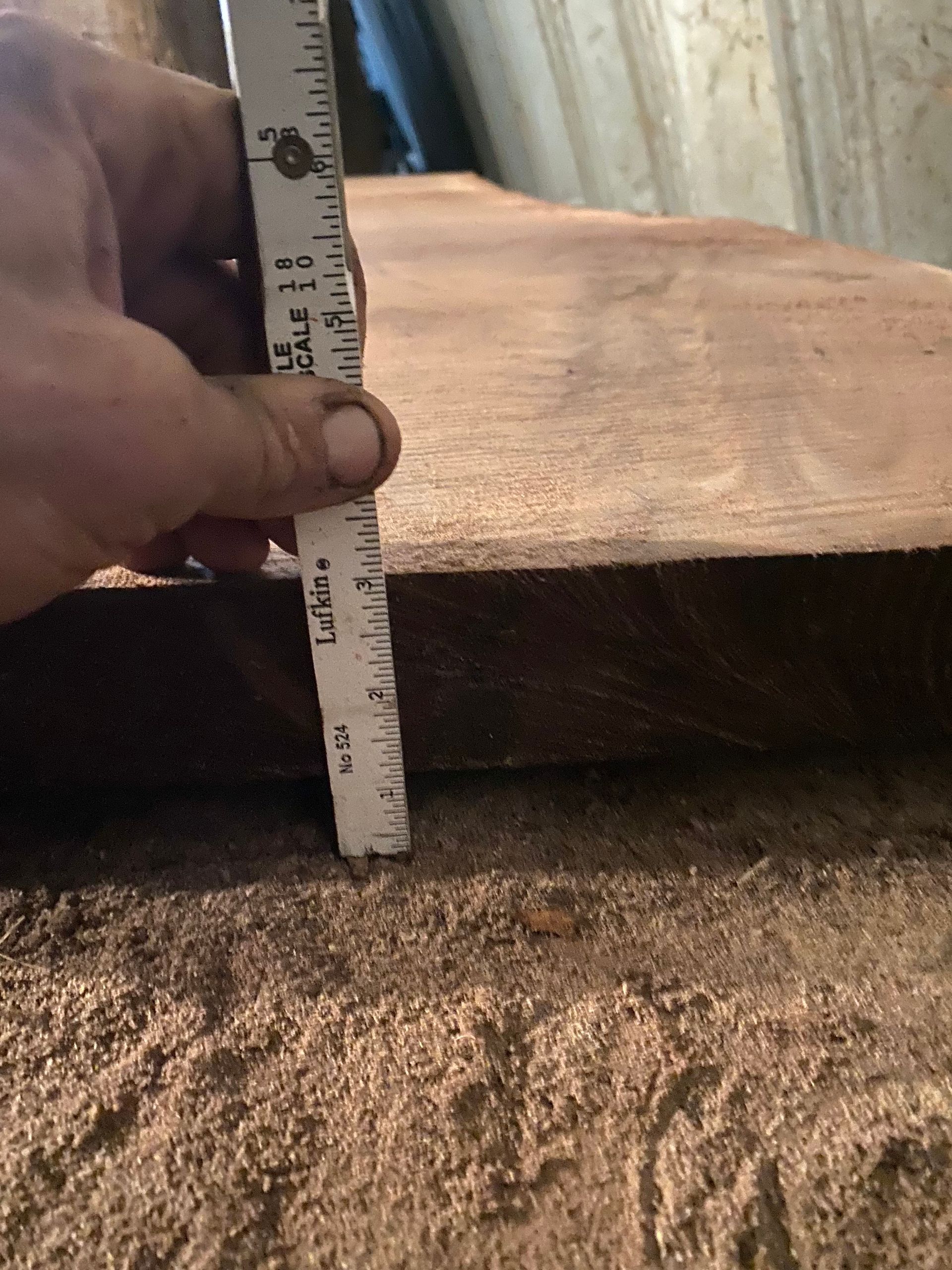 Hand holding ruler measuring thickness of a brown wooden board. Sawdust covers the floor.