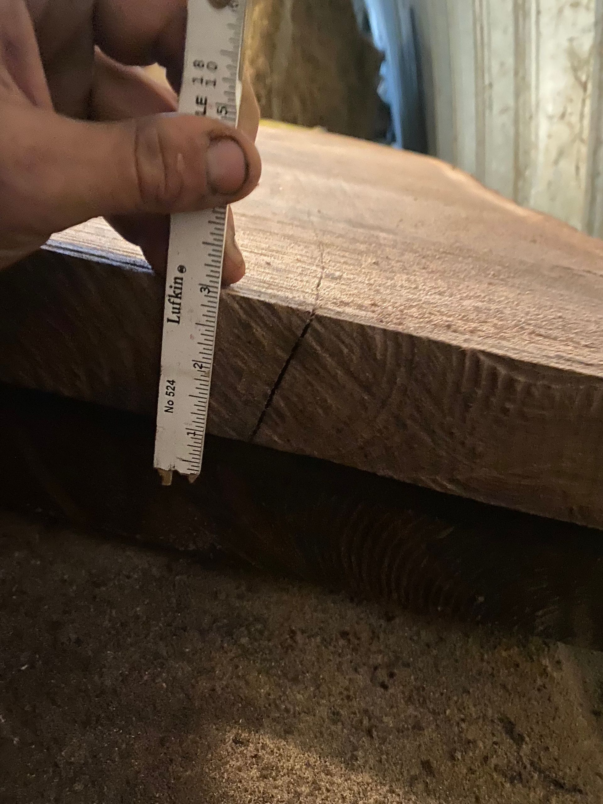 Hand holding a ruler, measuring the thickness of a wooden plank with a visible cut line.