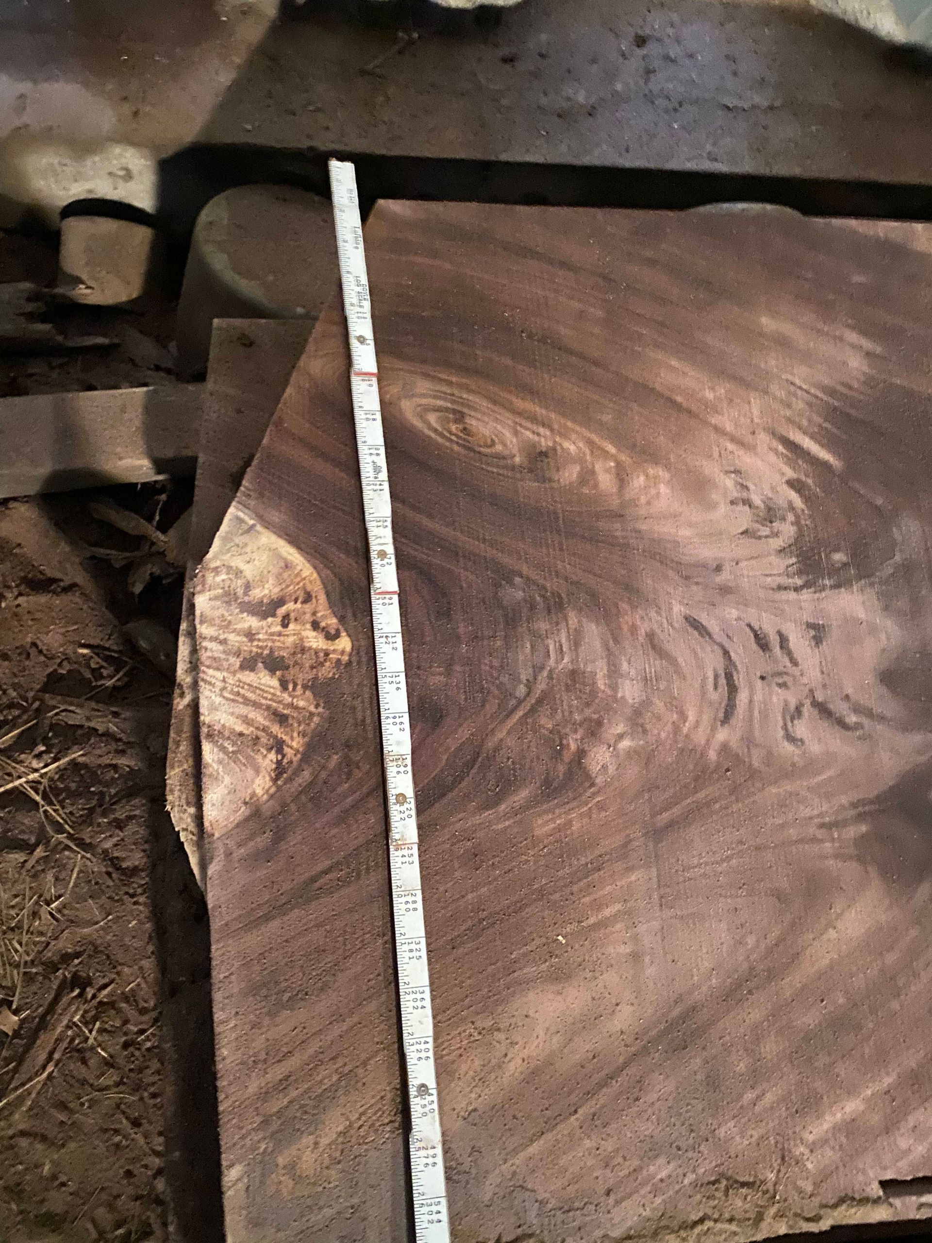 A wooden slab with a measuring tape laid across it, showing dark brown wood grain.