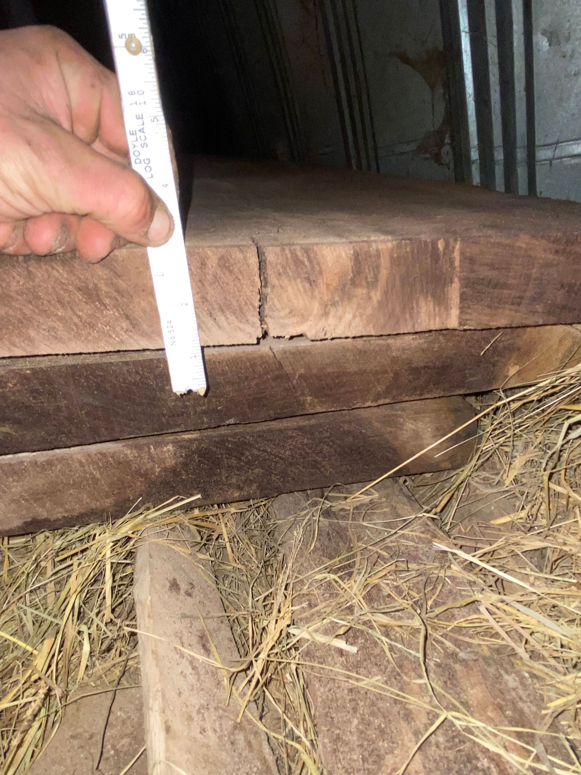 Hand holding a ruler next to stacked dark wooden planks with a crack, in a dim setting.