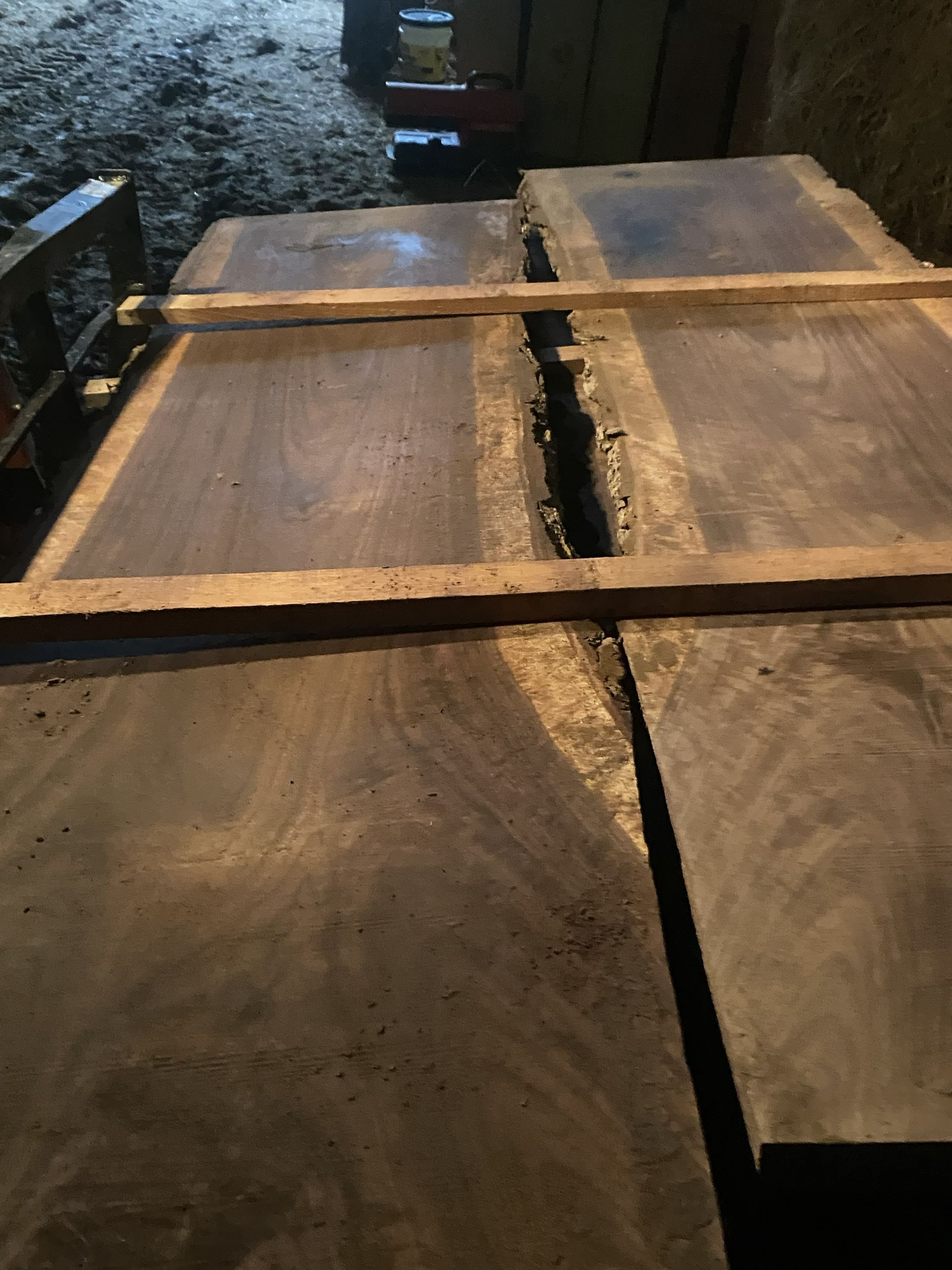 Dark wooden planks stacked, with a split and wood supports, in a dimly lit setting.