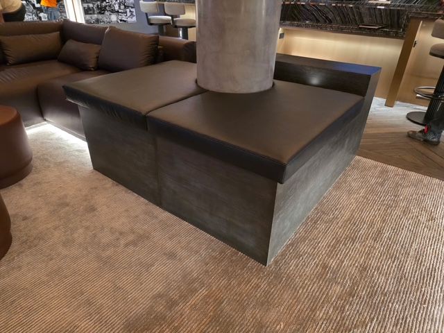 Gray wooden built-in seating surrounds a pillar in a modern lounge. Dark gray cushions sit on top.