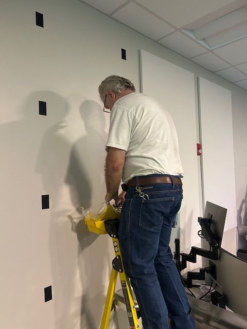 Man on a ladder working on a white wall with black squares. Indoor setting.