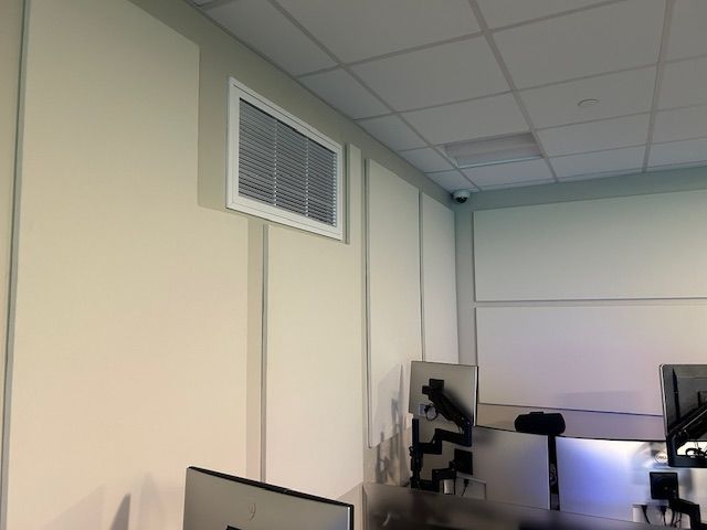 An air vent on a beige wall in a room with a white ceiling and monitors.