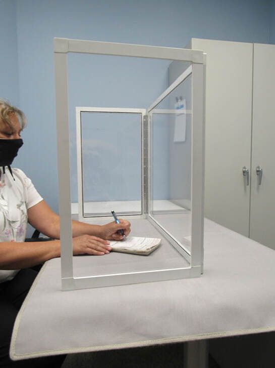 A woman wearing a mask is sitting at a desk behind a clear barrier.