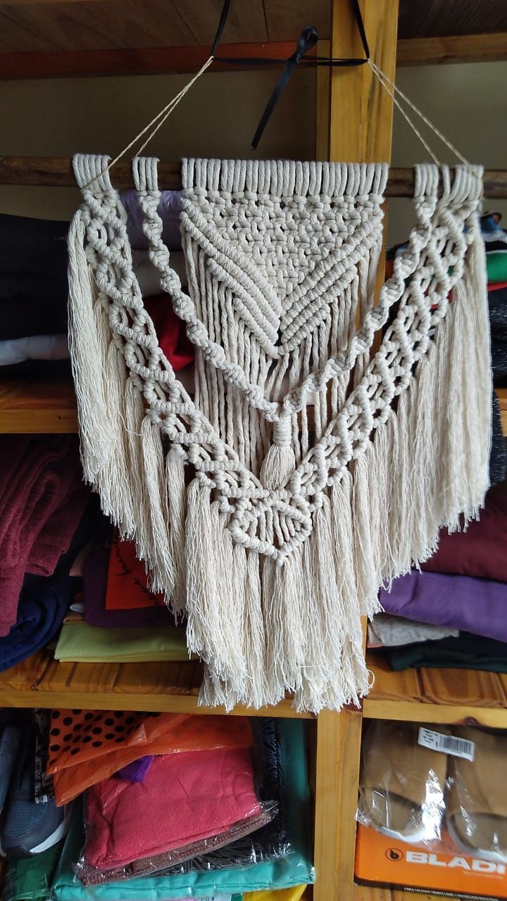 Tapiz de macramé blanco con flecos triangulares, expuesto en la estantería de una tienda.