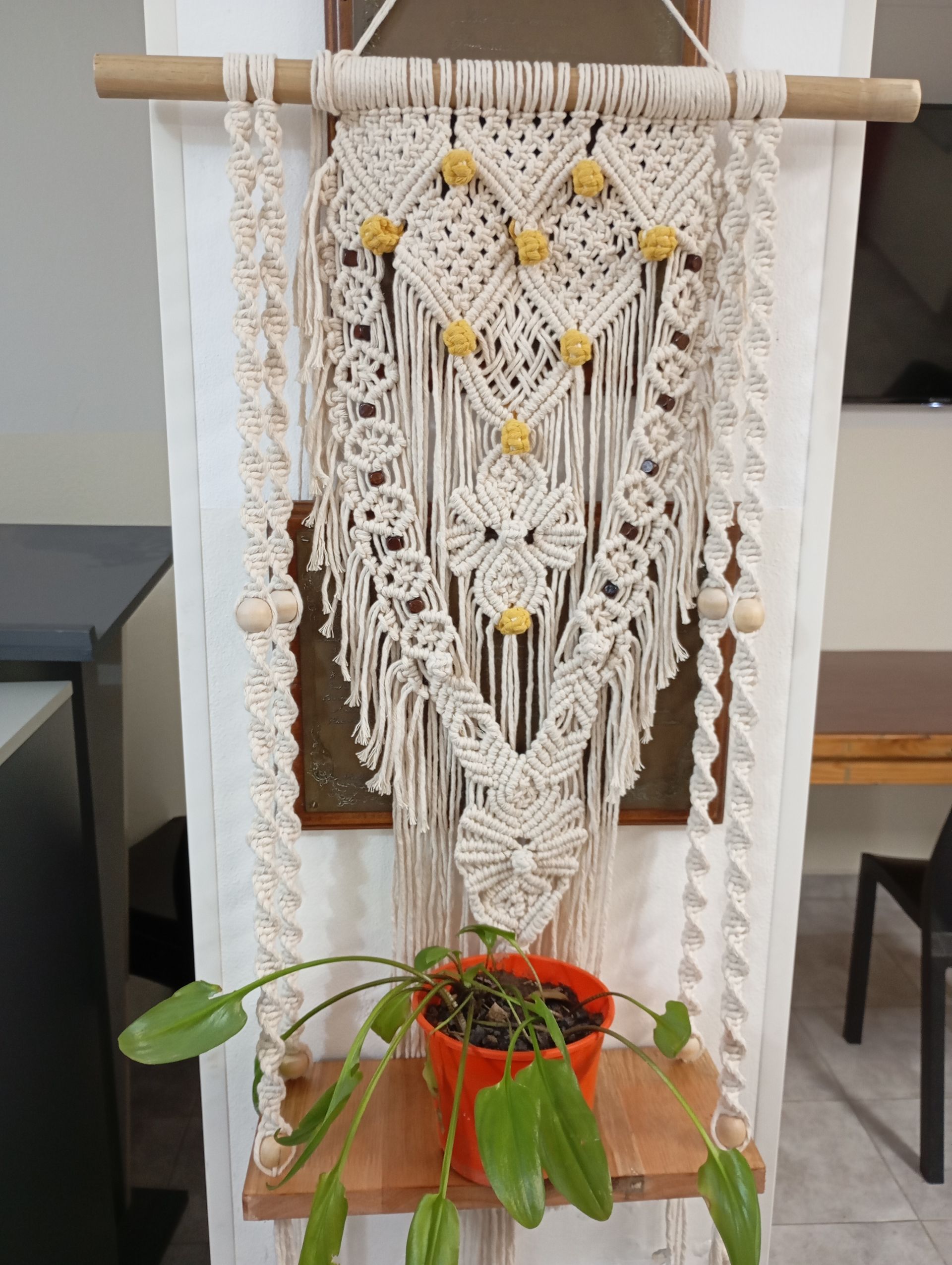Tapiz de macramé con cordones color crema, detalles amarillos y una planta colgante frente a una ventana.