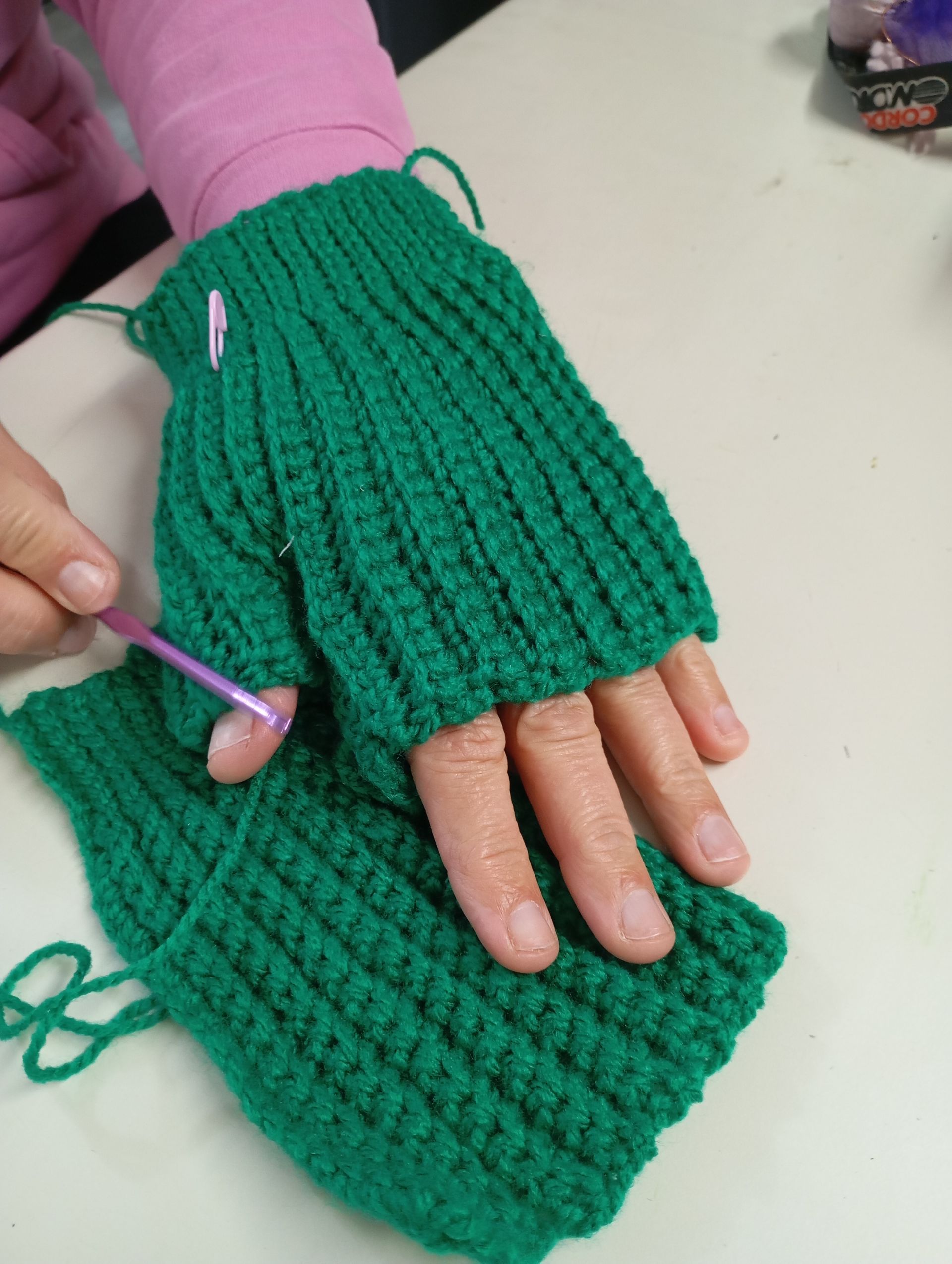 Manos con guantes verdes sin dedos tejidos a crochet, mientras que una de ellas está siendo cosida con un ganchillo rosa.