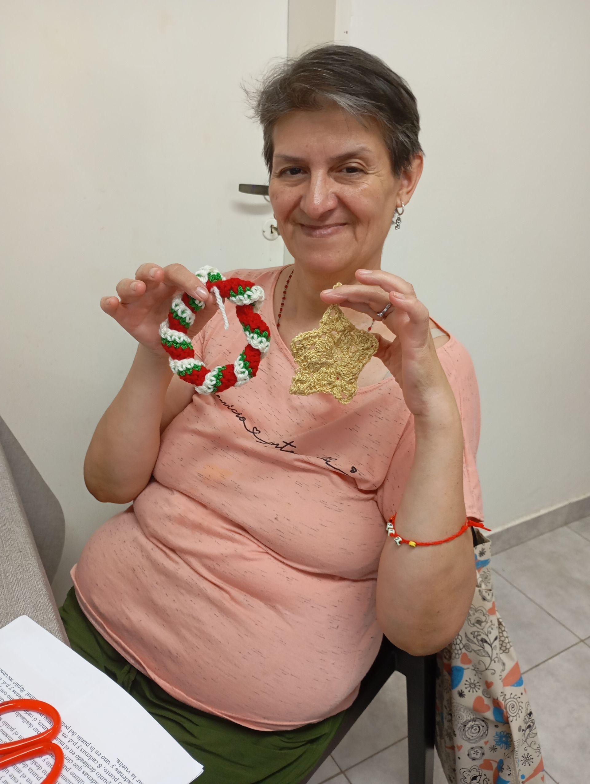 Mujer sentada en el interior de una vivienda, sosteniendo una corona roja y verde y un adorno dorado, sonriendo a la cámara.