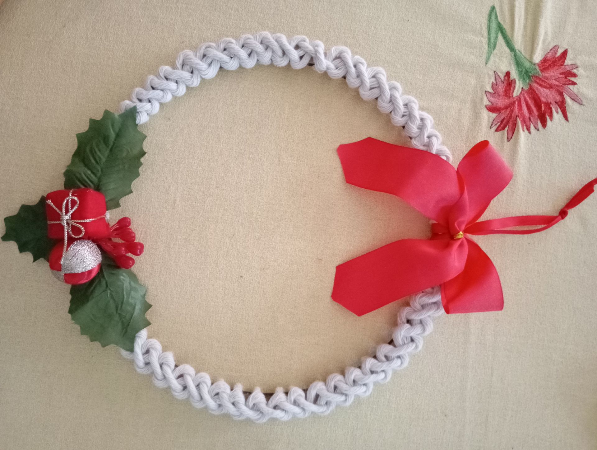 Corona navideña blanca trenzada con cinta roja, hojas de acebo, bayas y un adorno de flor roja sobre fondo beige.