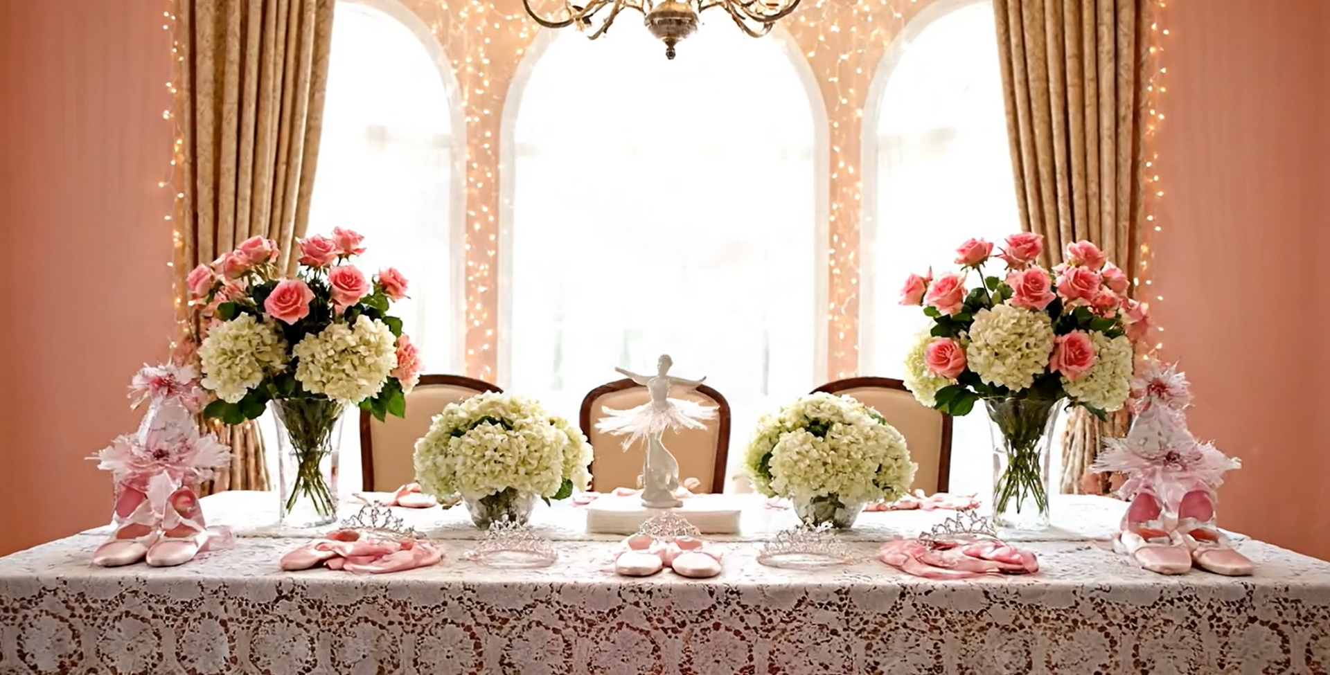 Table set for a celebration, featuring floral centerpieces, ballet shoes, and decorations in a light-filled room.