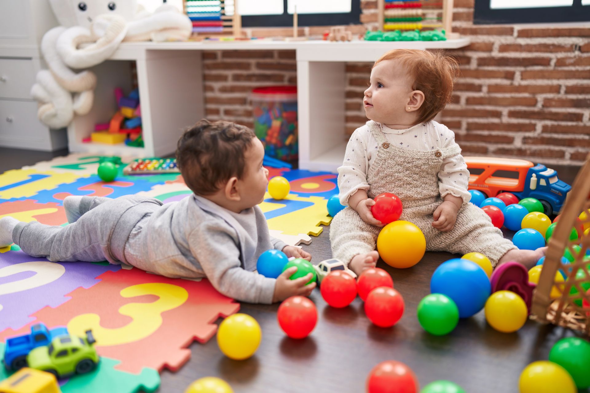Dos bebés jugando con pelotas de colores sobre una alfombra de espuma en una sala de juegos.