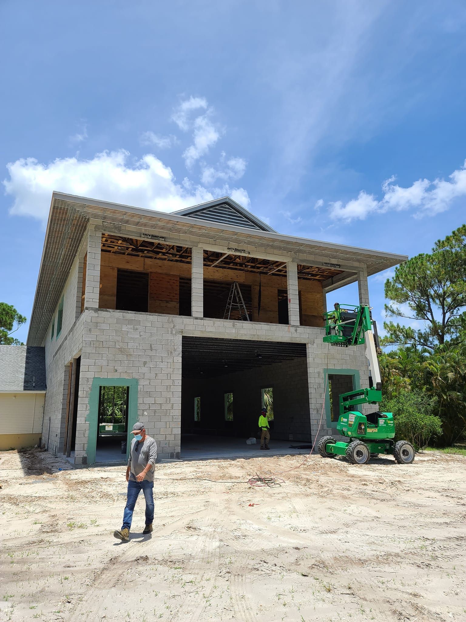 Construction site. Two-story building with stone facade, unfinished, man standing, and green machinery under blue sky.