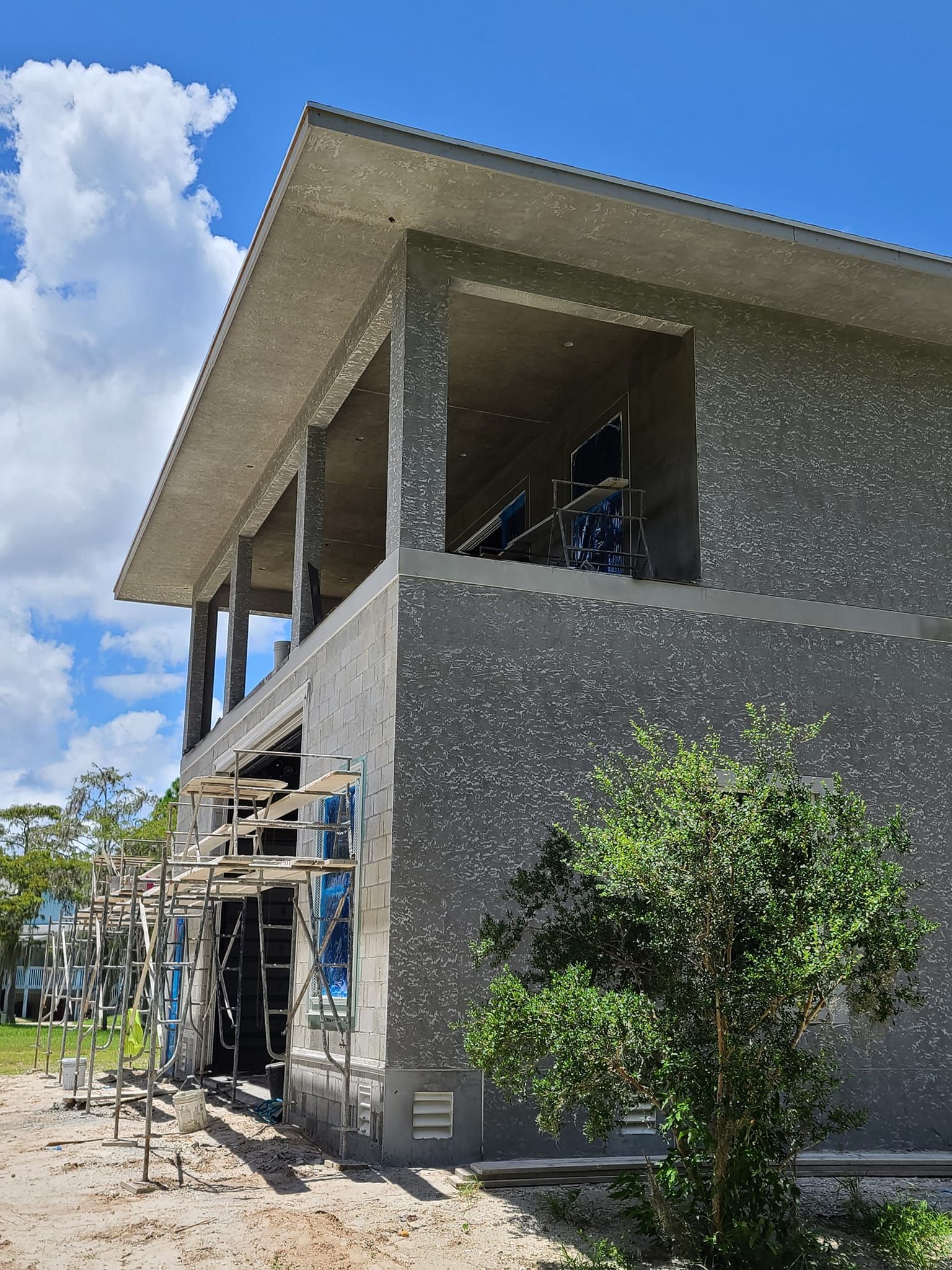 Building under construction; gray stucco walls, blue sky, green shrub, and empty window frames.