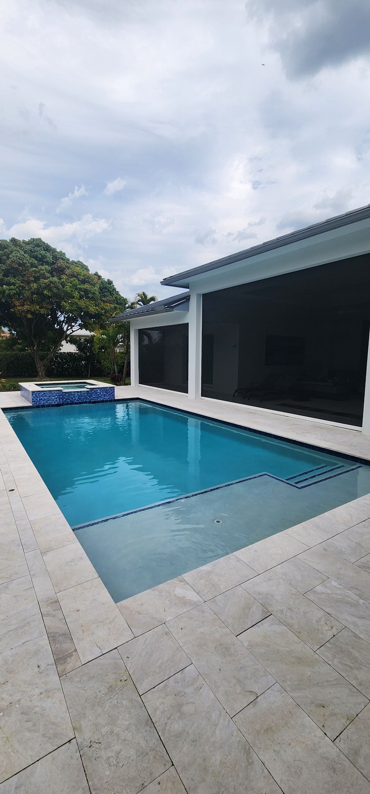 Pool with turquoise water, a patio, and a covered outdoor area under a cloudy sky.