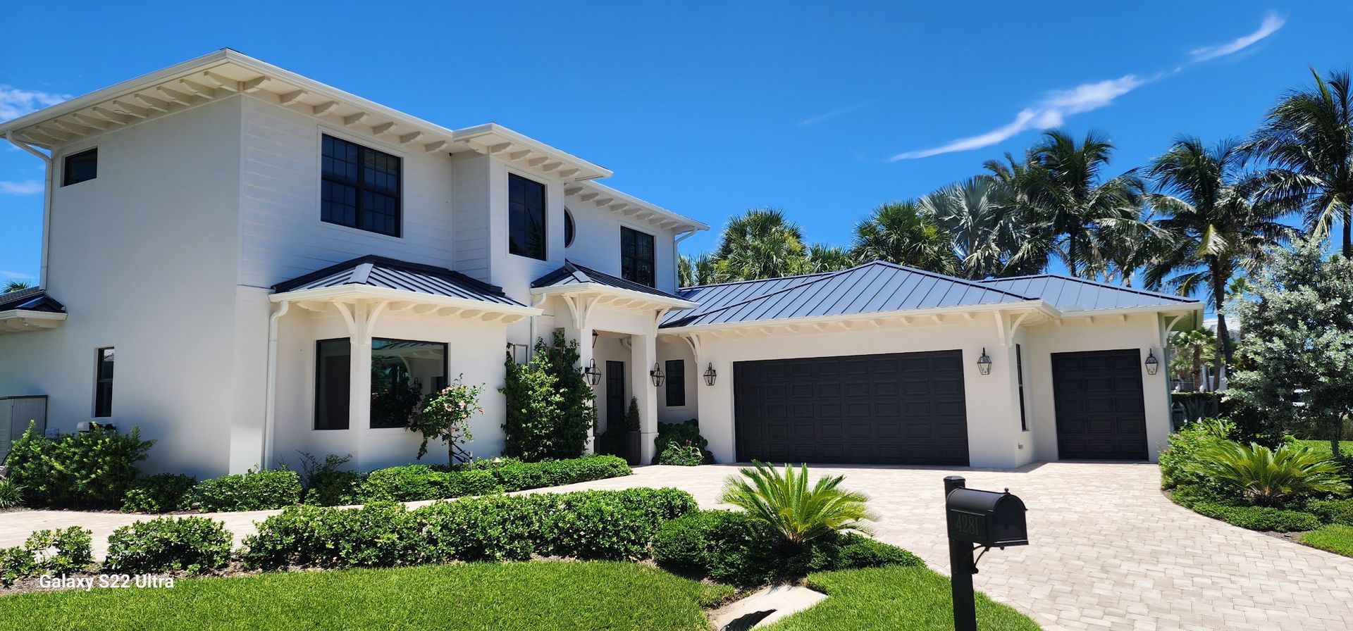 White two-story house with dark roof and garage doors, surrounded by green landscaping and a blue sky.