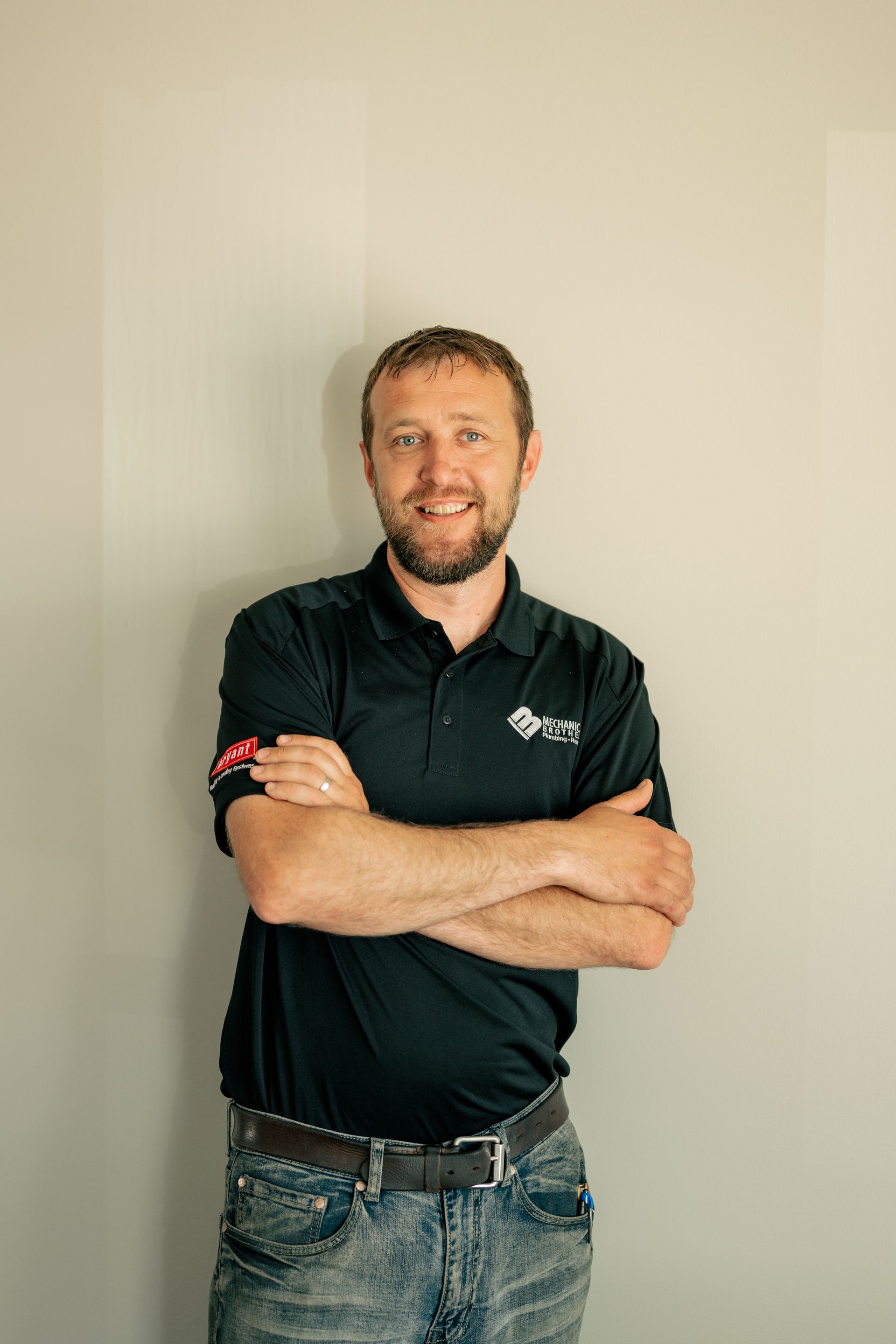 A man with a beard is standing with his arms crossed in front of a wall.