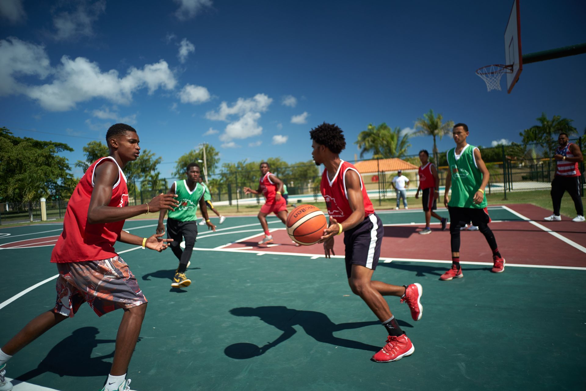 Basketball players in red and green jerseys on an outdoor court under a blue sky