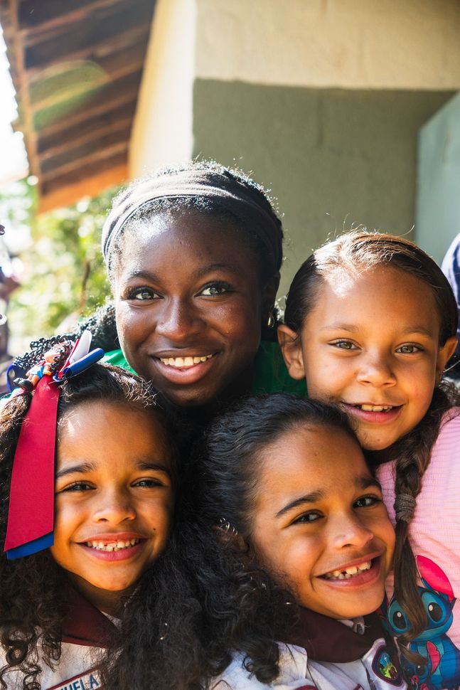 Four smiling children posing closely together outdoors near a building with sunlight and colorful clothing.