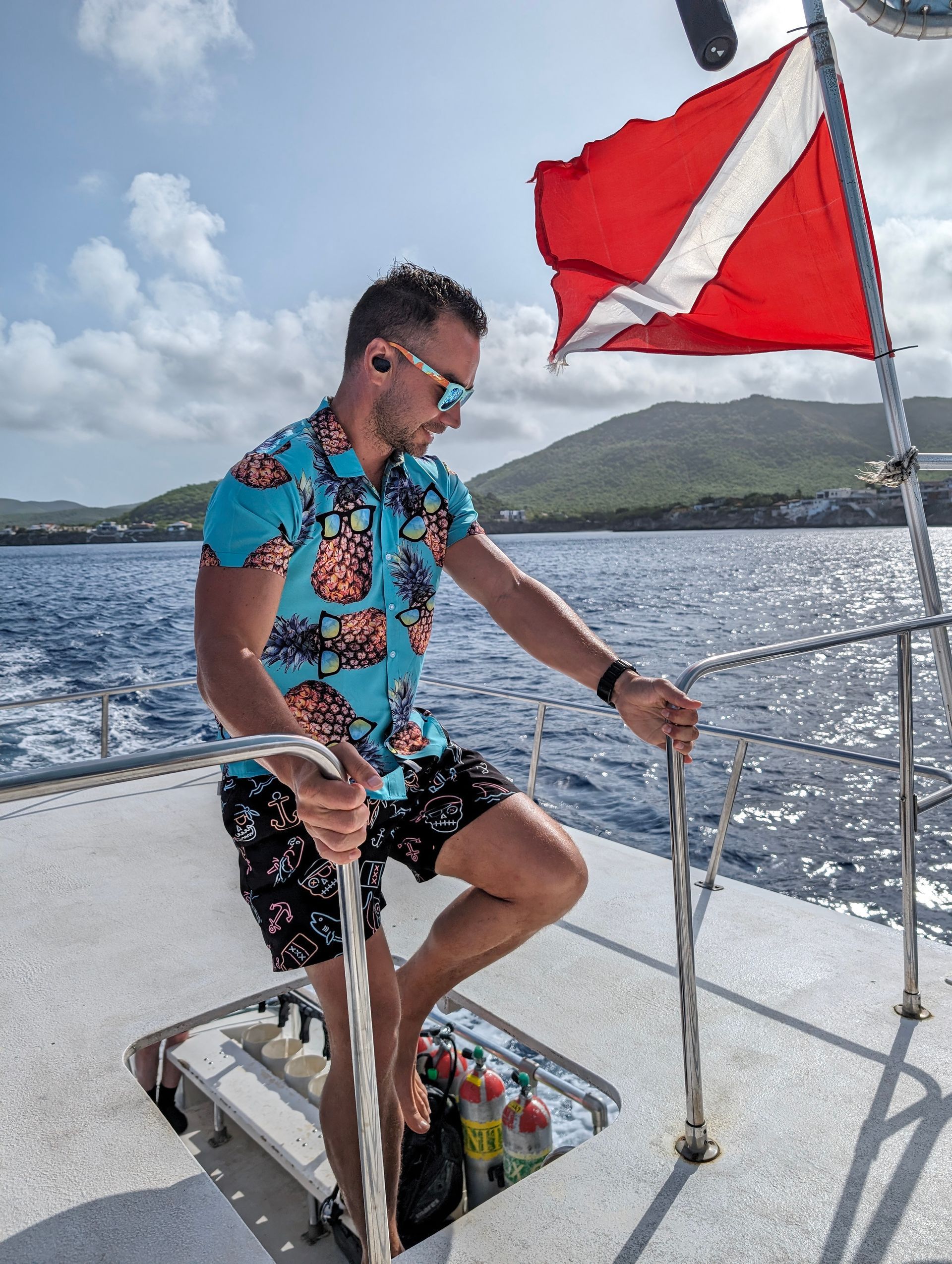 Man in floral shirt, shorts, sunglasses on a dive boat; red and white dive flag.