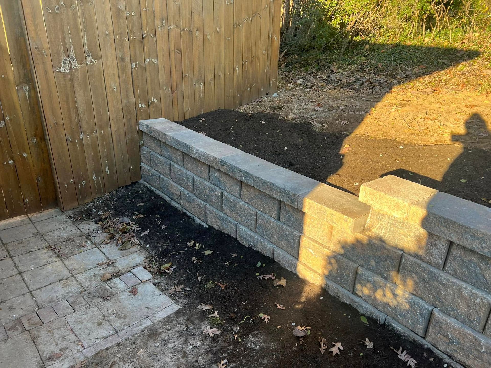 Backyard corner with a low concrete block wall, wooden fence, and paved patio in sunlight