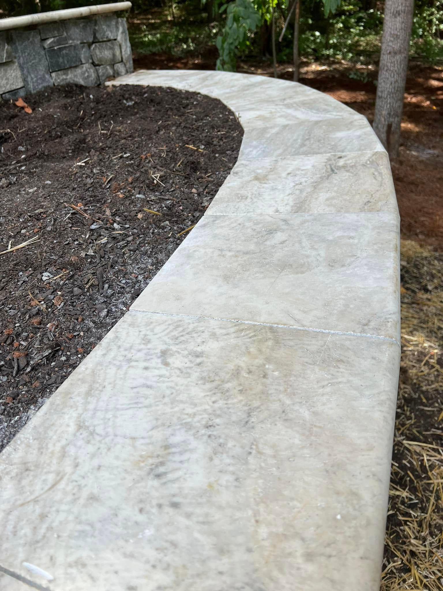 Curved light-gray concrete walkway beside a mulched garden bed and stone wall outdoors