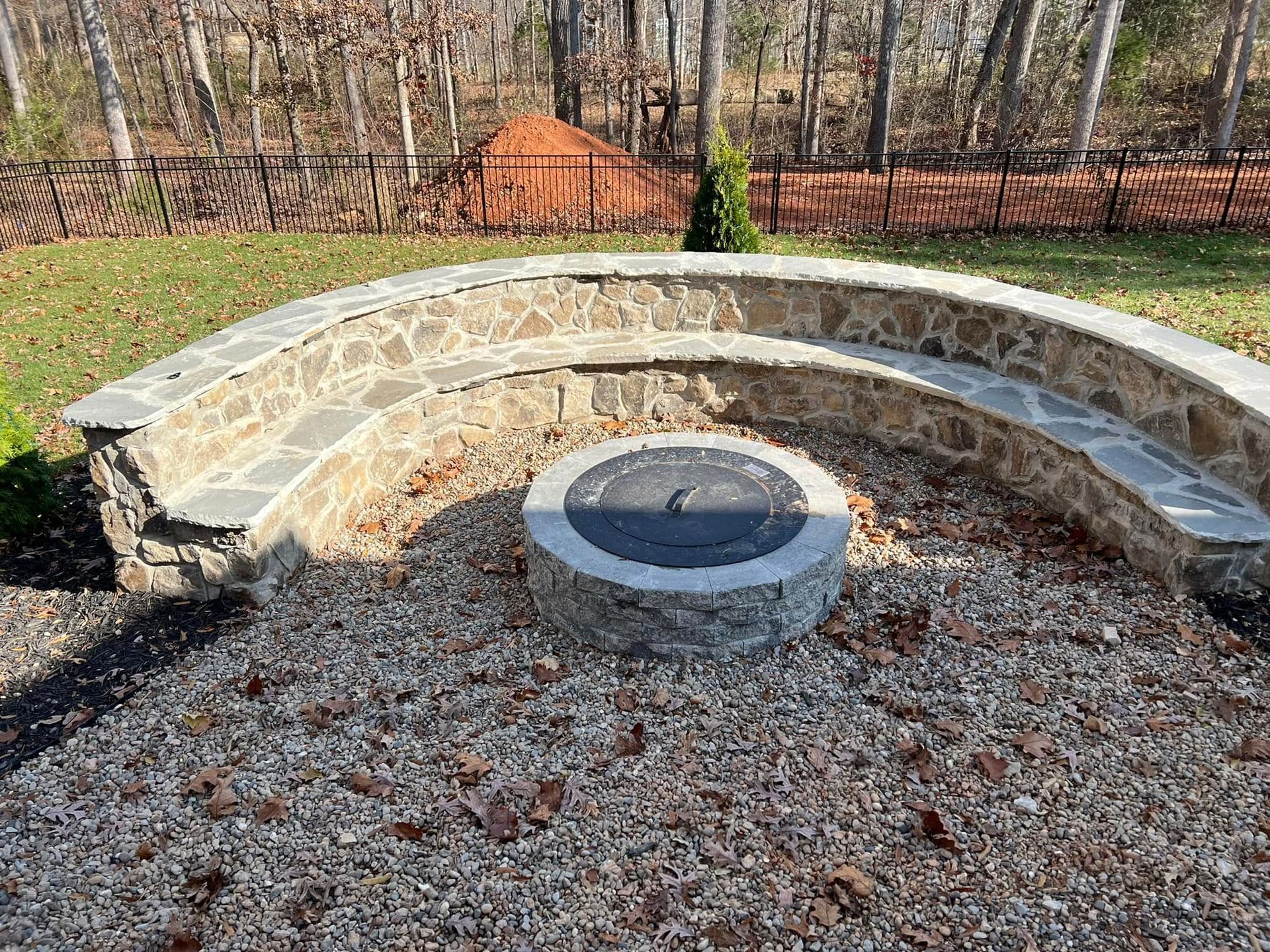 Stone semicircular bench around a circular fire pit in a leaf-covered outdoor area.