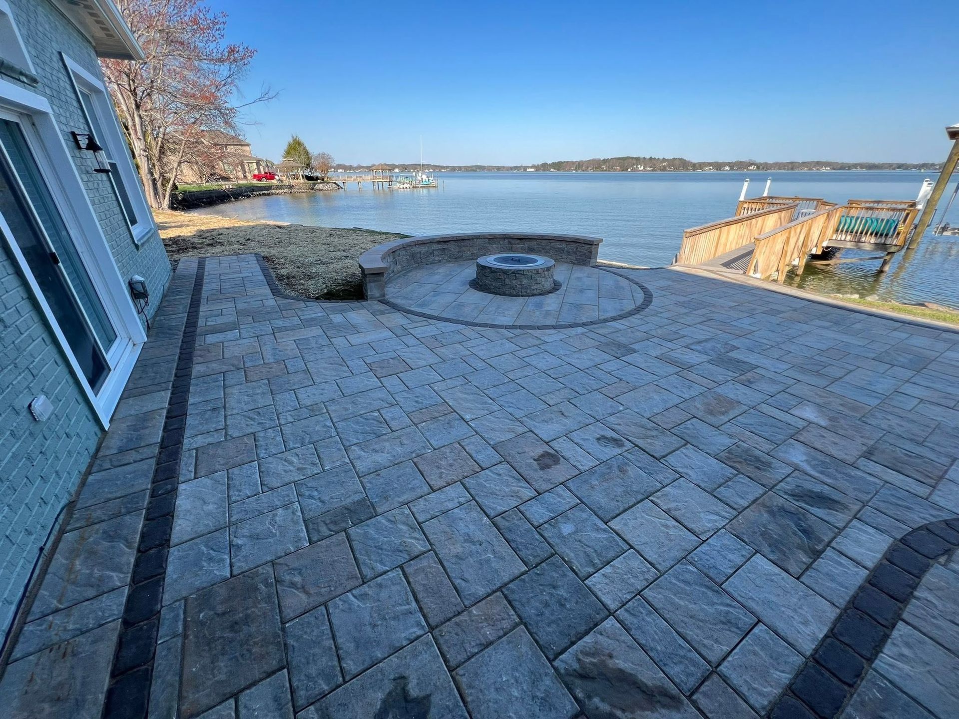 Stone patio overlooking a calm lake on a clear day, with a round fire pit and blue house wall.