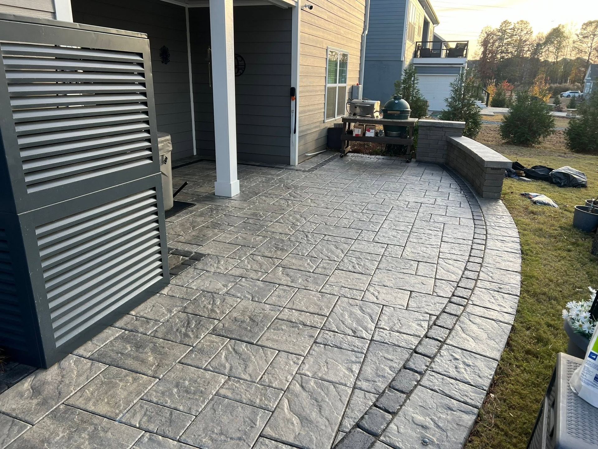 Curved stone patio beside a house with a covered seating area and grill, bordered by lawn and trees