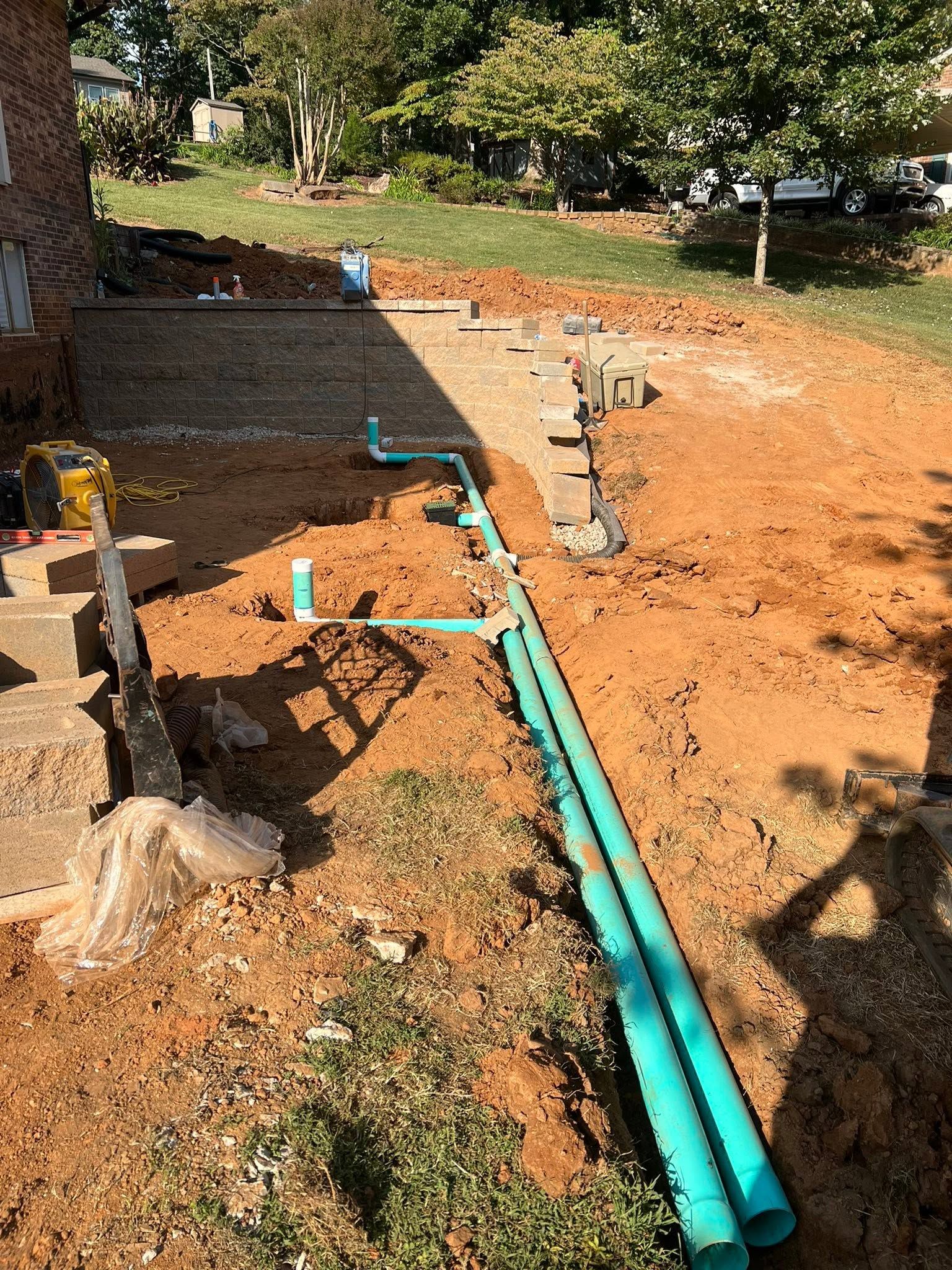 Construction site with turquoise pipes laid across dirt beside a retaining wall and steps under trees