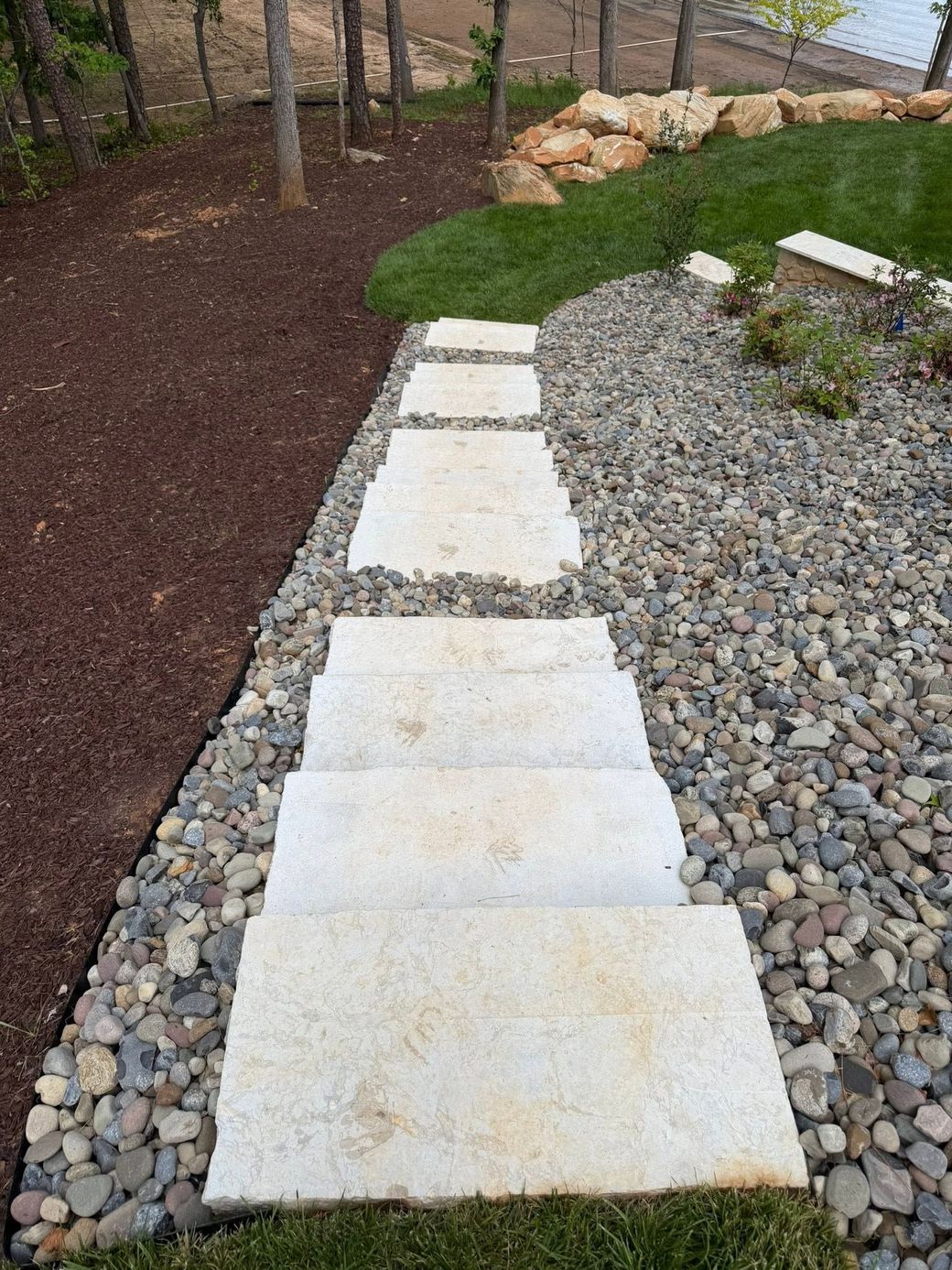Stone stepping path through a landscaped garden with gravel, mulch, trees, and a rock border