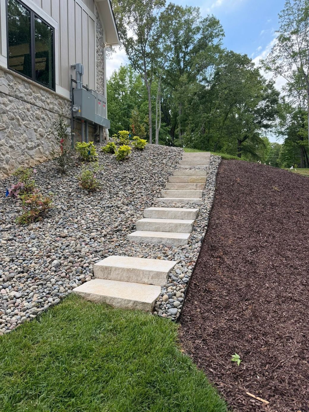 Stone steps up a landscaped slope beside a building, bordered by gray and burgundy gravel and green grass.