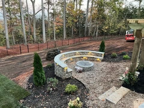 Backyard patio with curved stone bench fire pit, gravel ground, and wooded yard beyond