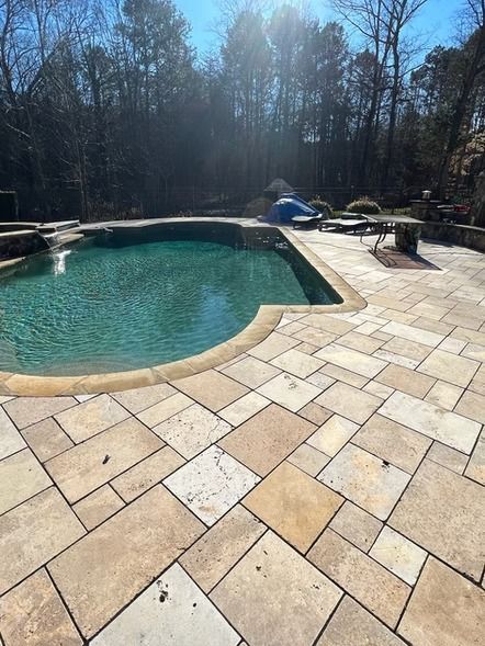 Backyard pool beside tan stone patio with lounge chairs and leafless trees in the background