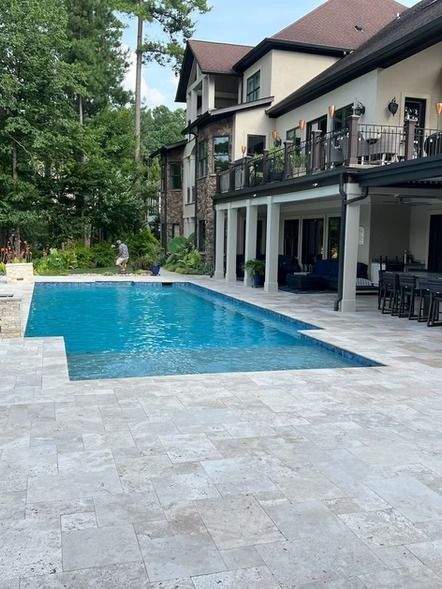 Modern backyard patio with a blue swimming pool beside a large house and trees