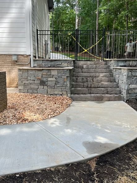 Stone steps and retaining wall beside a curved concrete walkway and house exterior, with a black fence above.