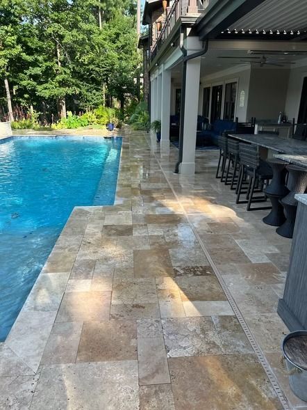 Poolside patio with stone tiles beside a blue swimming pool and shaded outdoor seating area