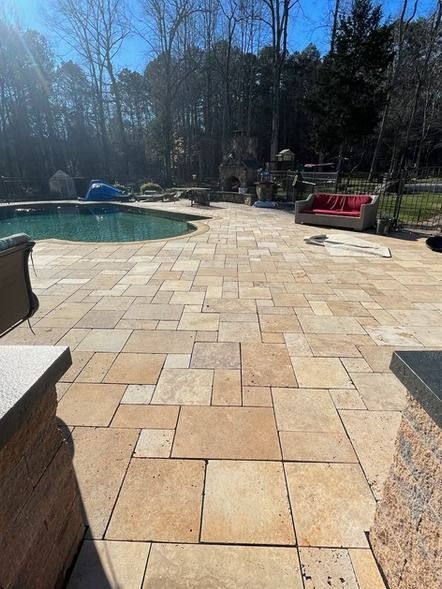 Backyard patio with tan stone pavers beside a curved pool, framed by trees and lounge chairs