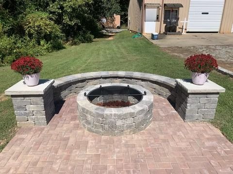Backyard stone fire pit with curved bench seating and potted red flowers on brick patio