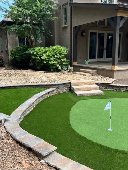 Small backyard putting green with a flagstick beside a stone patio and steps to a house