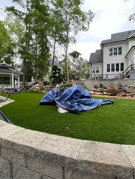 Blue tarp-covered object on a manicured lawn in front of a large house and trees