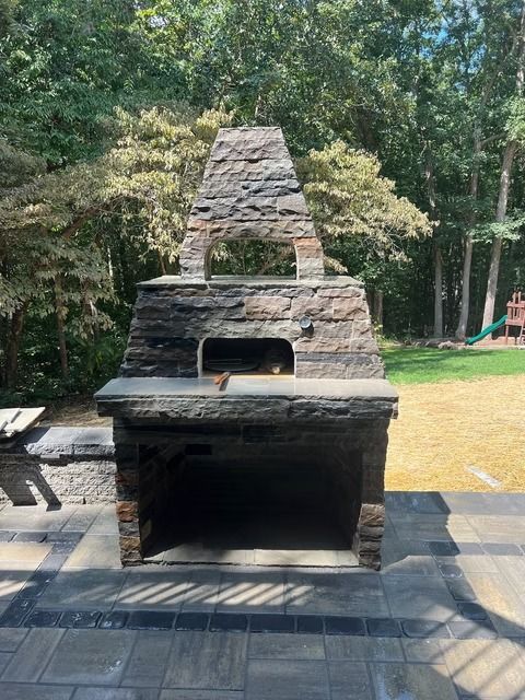 Stone outdoor fireplace with a tall chimney on a patio, surrounded by trees and grass.