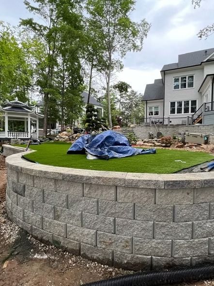 Curved stone retaining wall around a landscaped lawn with a blue tarp, near a house and gazebo