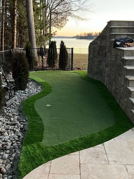 Curved artificial turf putting green beside a stone patio, with rocks, trees, and a lake at sunset.