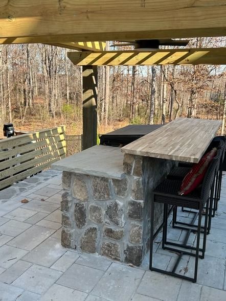 Stone outdoor bar under a wooden pergola with stools and a tiled patio, surrounded by trees.