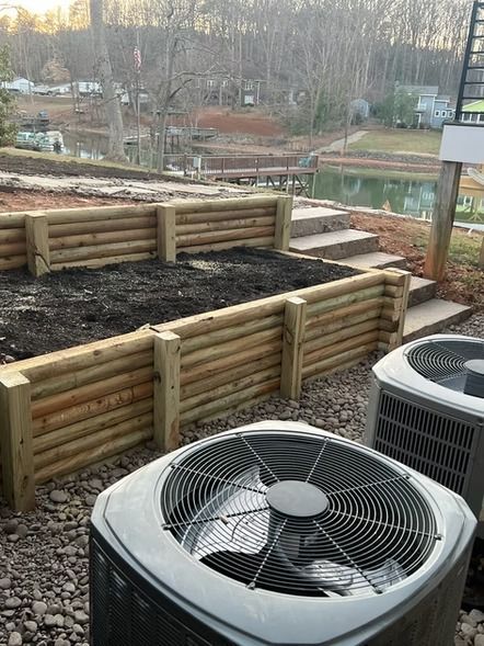 Raised wooden garden beds beside two air-conditioning units in a backyard, with bare trees in the background
