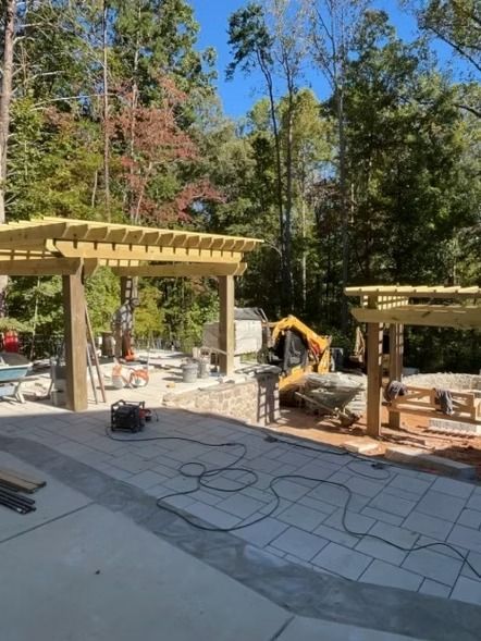 Outdoor patio construction with wooden pergolas, concrete slab, and an excavator in a wooded yard